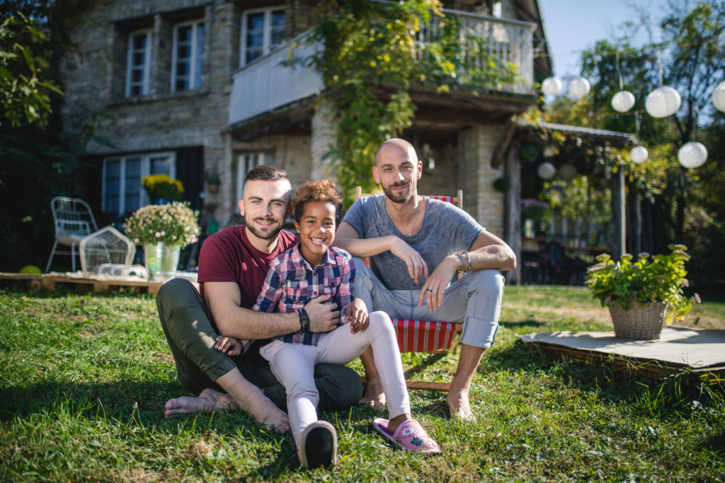 Same-sex adoption: two fathers sit on the grass outside their home with their smiling young son between them