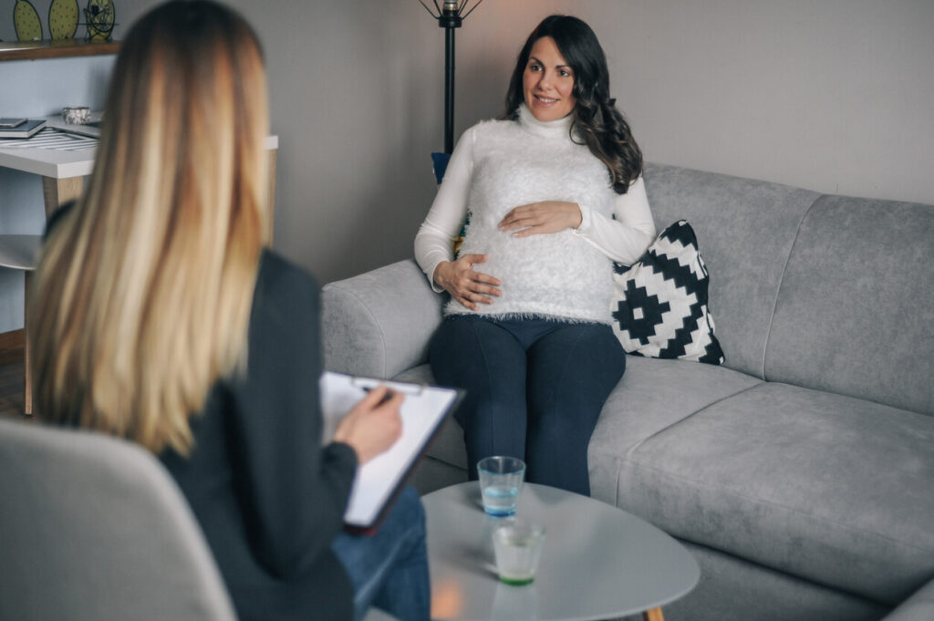Independent social worker assessment at home: a professional takes notes while talking with an expectant mother on her sofa