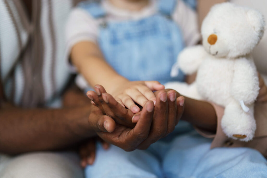 Post adoption contact: an adult's hands holding a young child's hands beside a white teddy bear